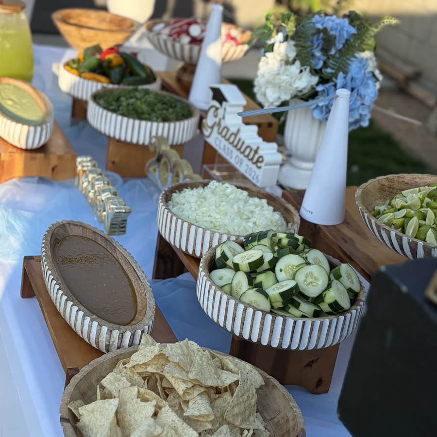 A graduation spread with ribbed bowls of red salsa, chopped onion, cucumber, lime and tortilla chips on wooden risers.