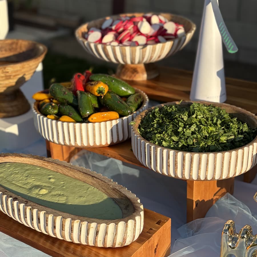 Fluted wooden bowls of sliced radishes, jalapeños, mini peppers, chopped cilantro and avocado salsa on wooden risers in late afternoon light.