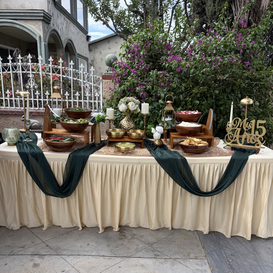 A quinceañera table set in cream linen with deep green draping and gold serving pieces.
