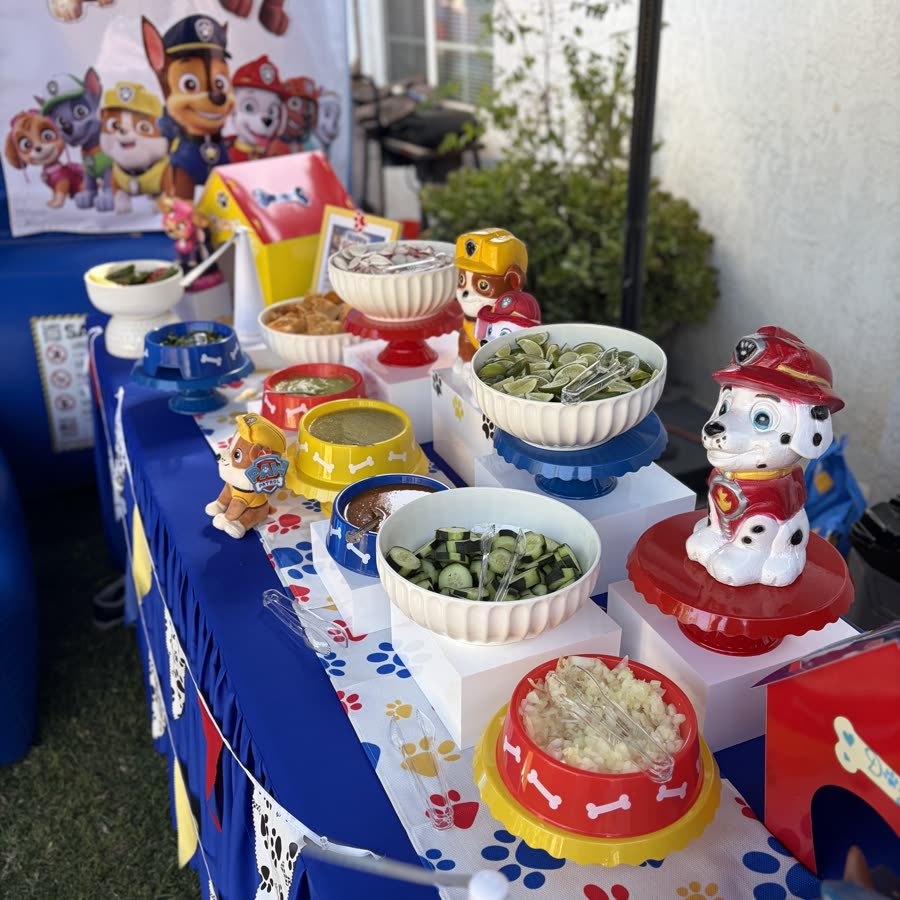 A Paw Patrol themed birthday table in blue, red and yellow, with dog bowl dishes of salsa, cucumber and lime on white risers.