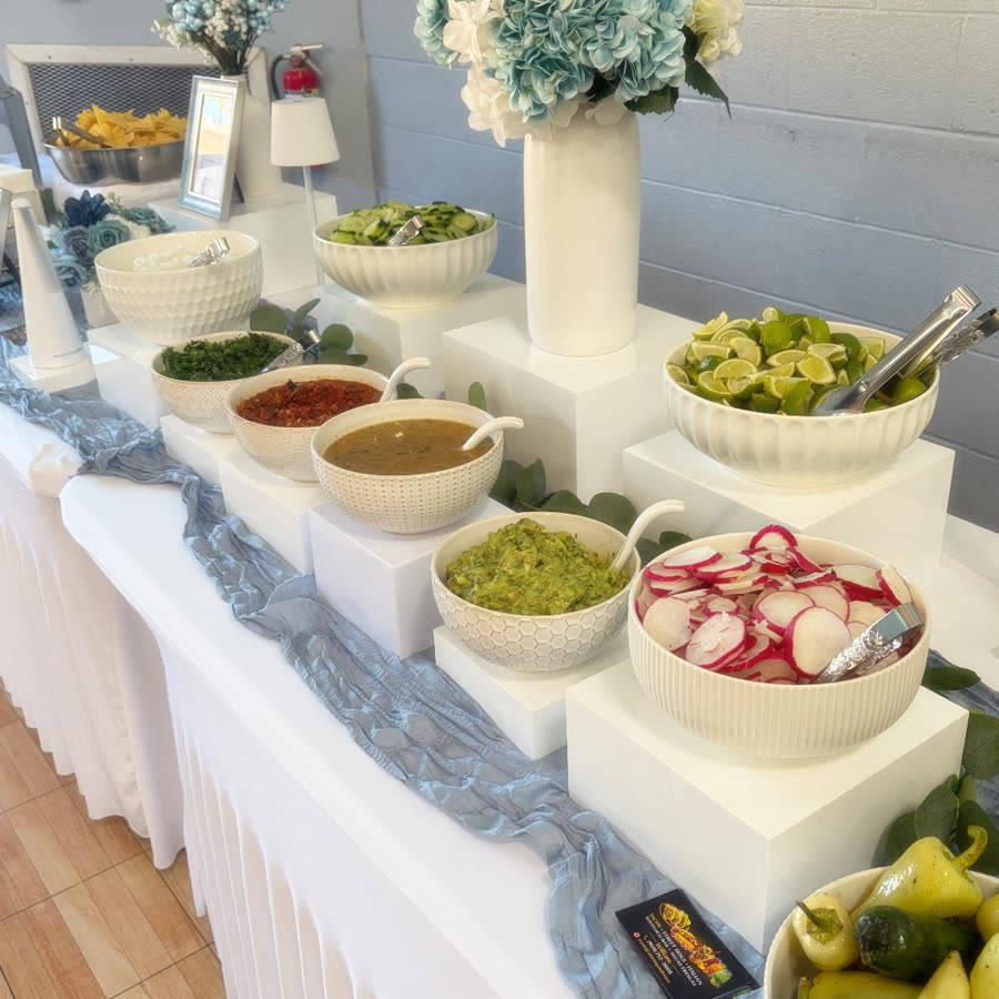 A salsa bar built on white risers with bowls of guacamole, radish, lime and cilantro, a dusty blue runner and pale hydrangeas.