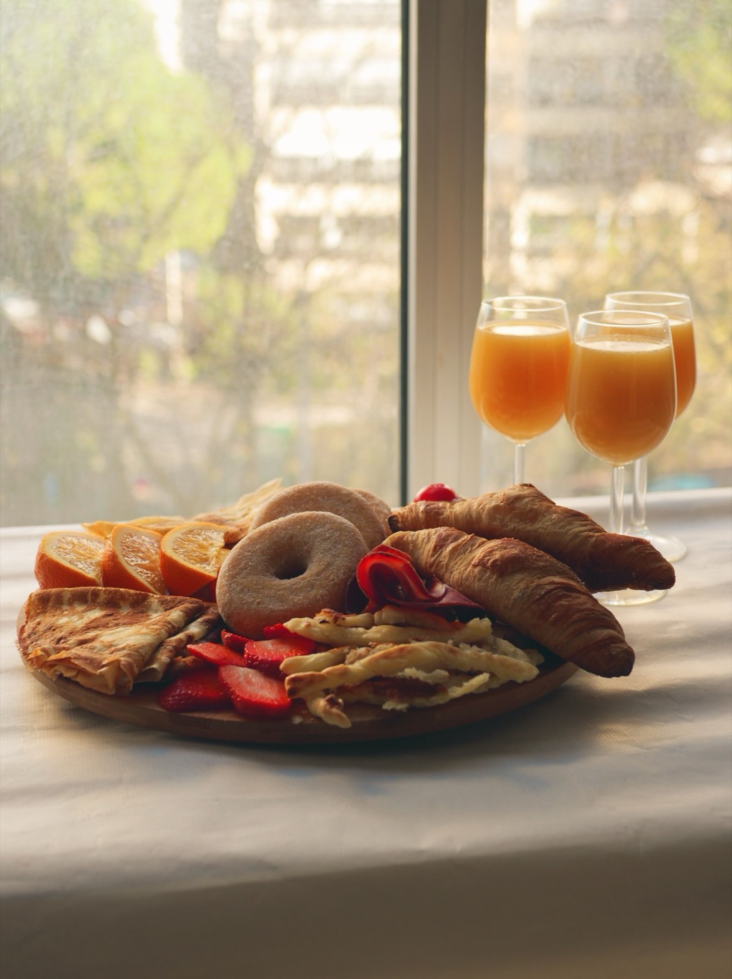 A brunch board of croissants, pancakes, a doughnut, strawberries and orange slices beside glasses of mimosa.